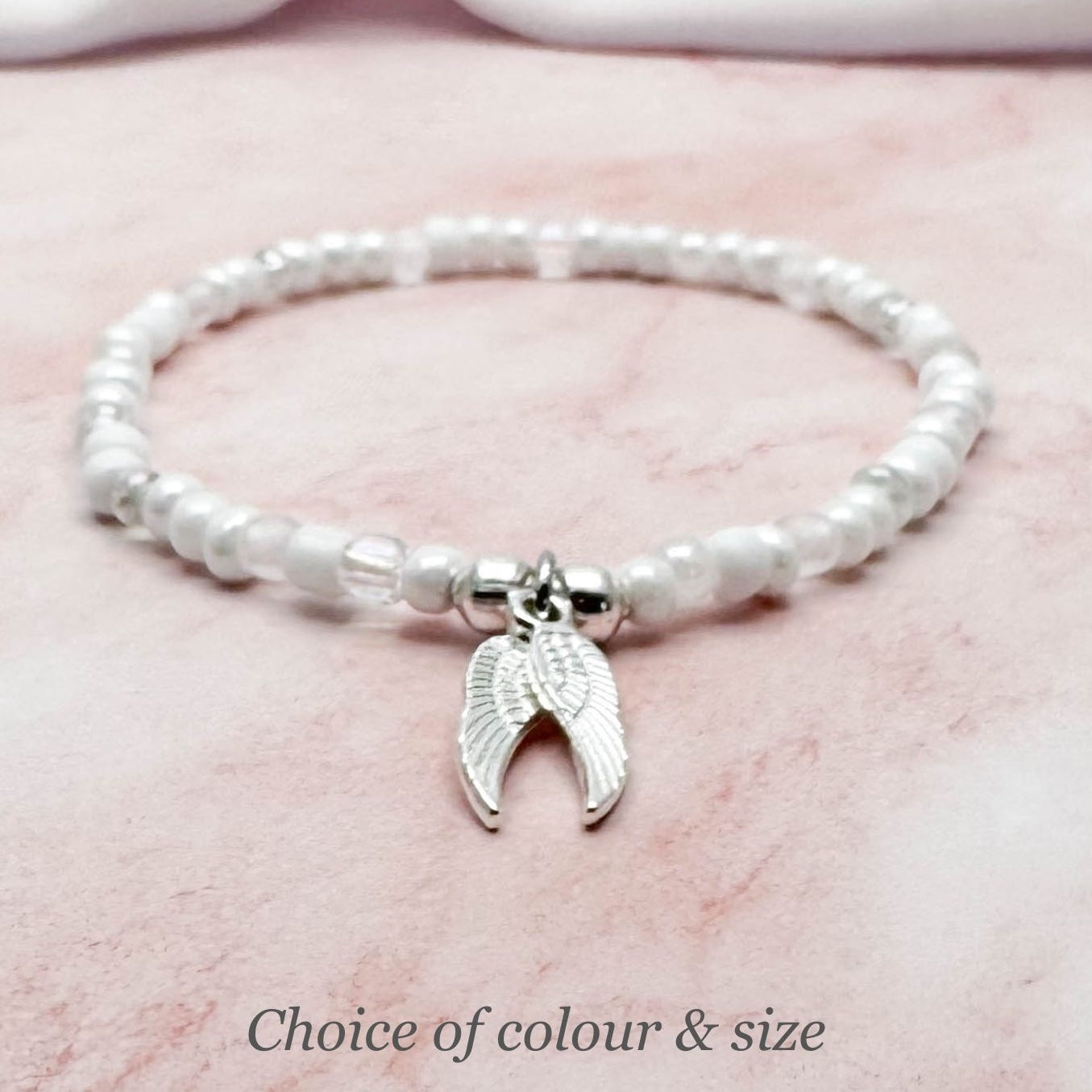 A white bead bracelet with a silver wing charm on a pink marble background.