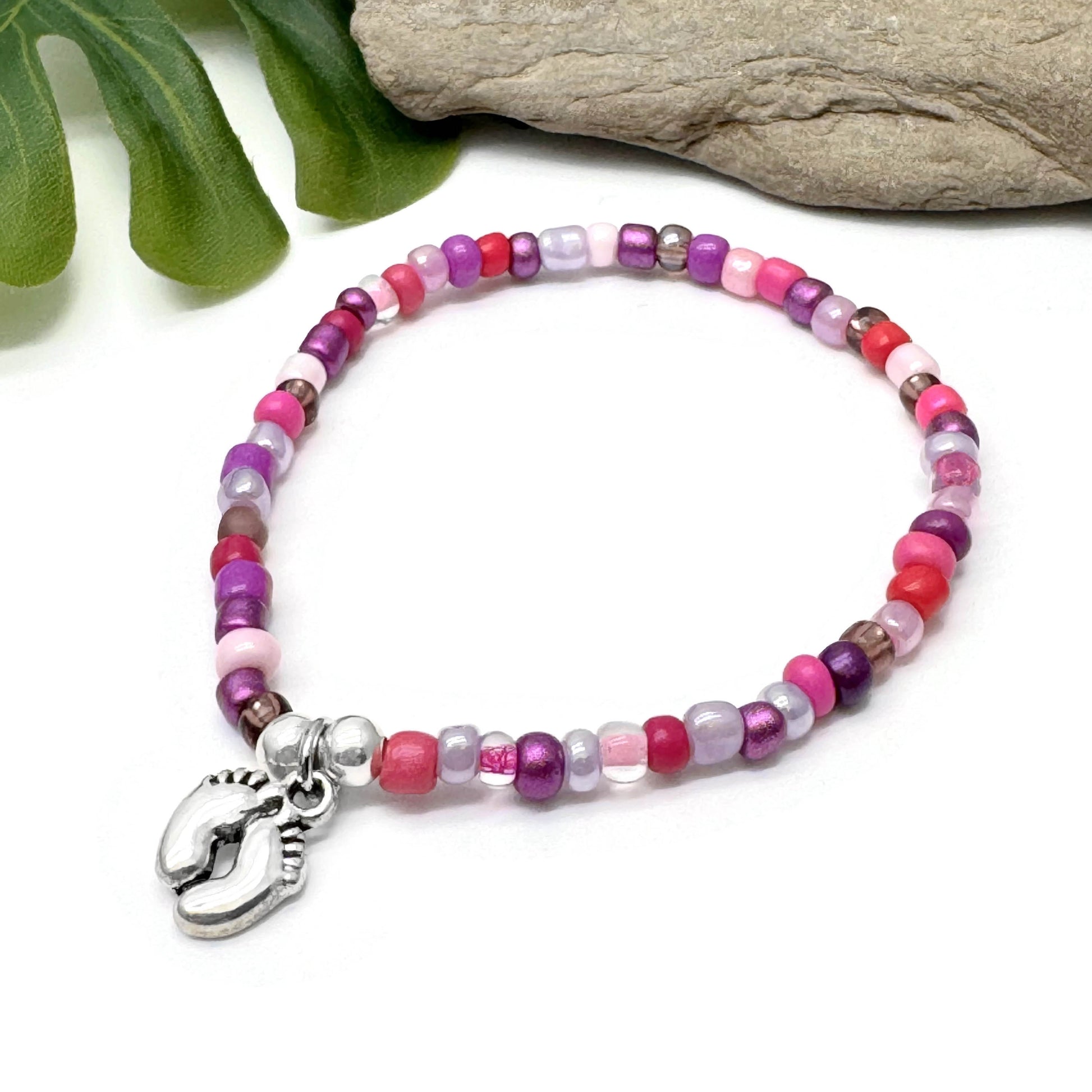 A colorful beaded bracelet with a charm featuring a pair of feet, placed on a white surface next to some green leaves.
