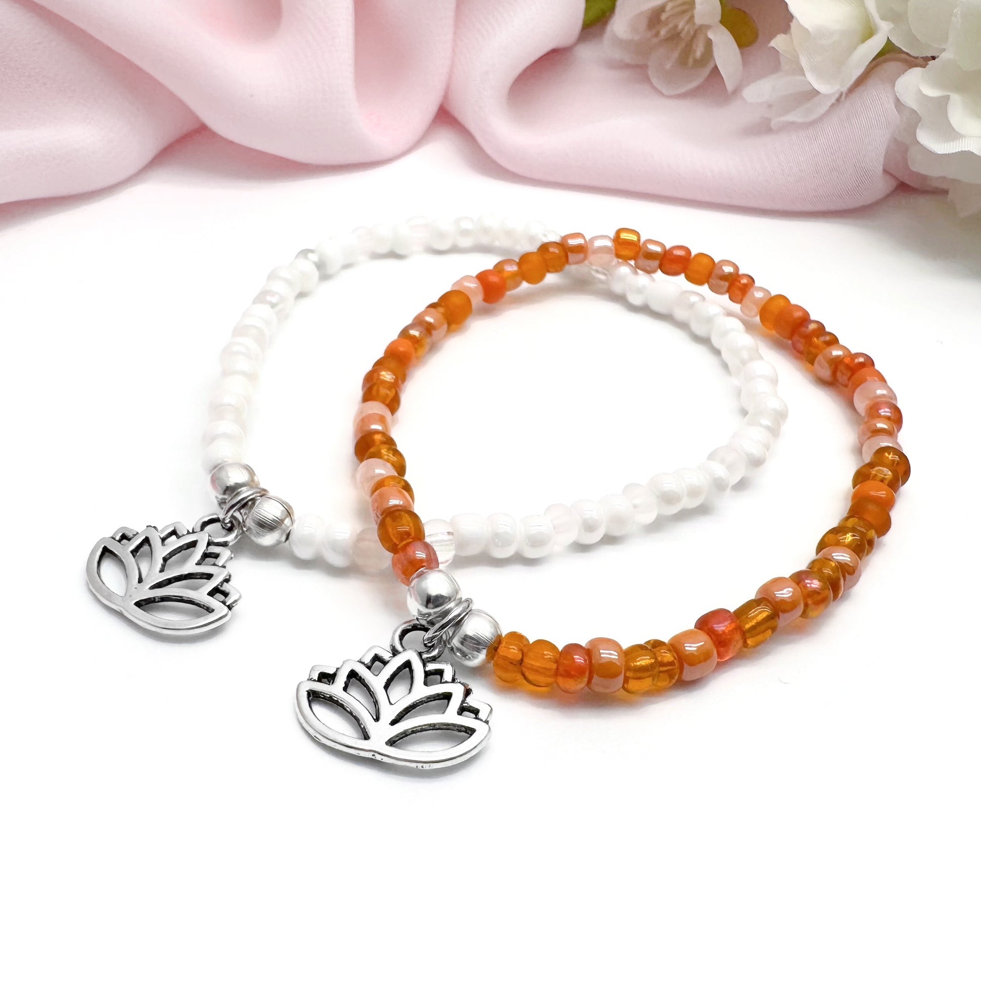 Two beaded bracelets with metal charms, one with a white bead and the other with an orange bead, placed on a white surface with a pink fabric background.