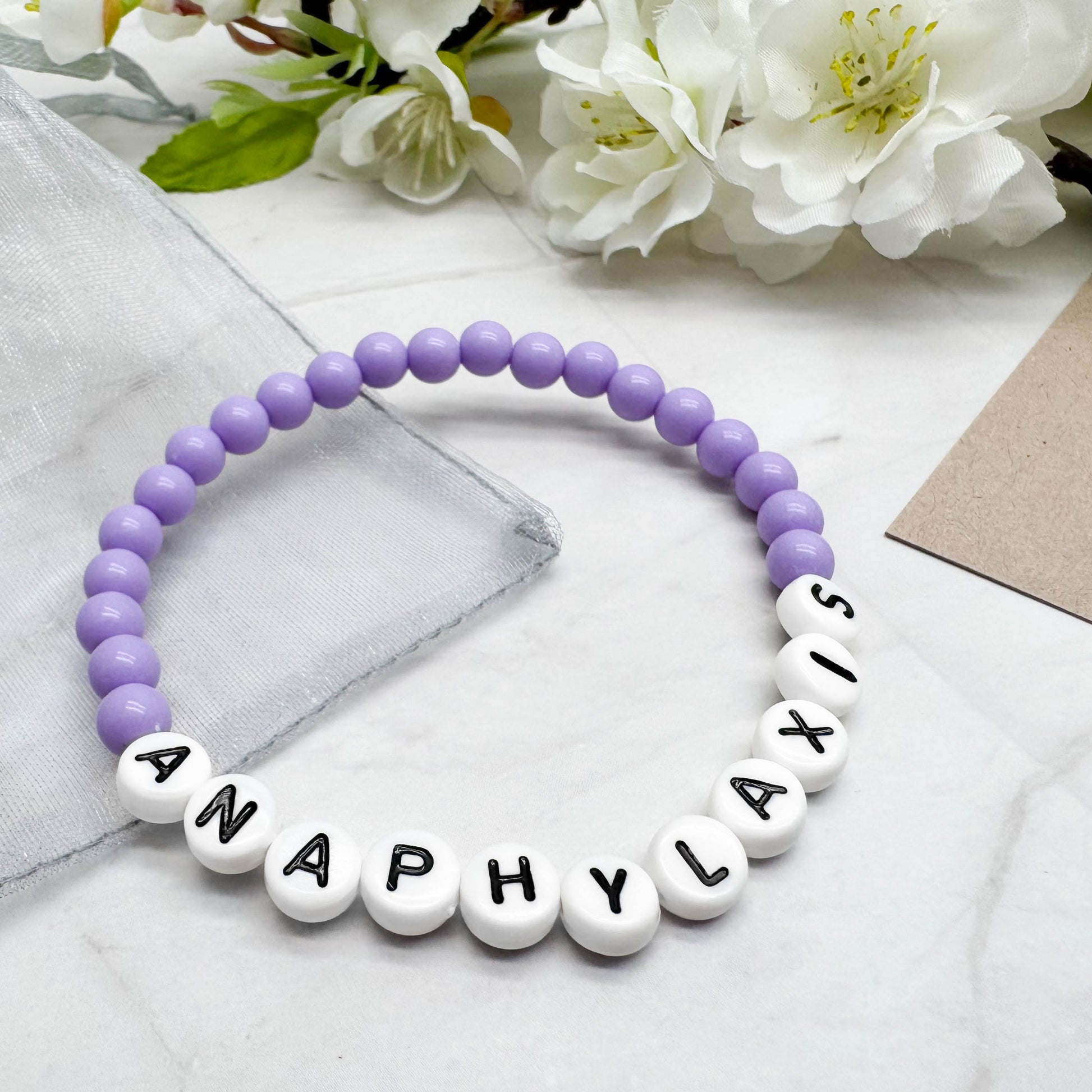 A bracelet made of purple beads and white beads spelling out the word "ANAPHYLaxis" is displayed on a marble surface, with white flowers in the background.