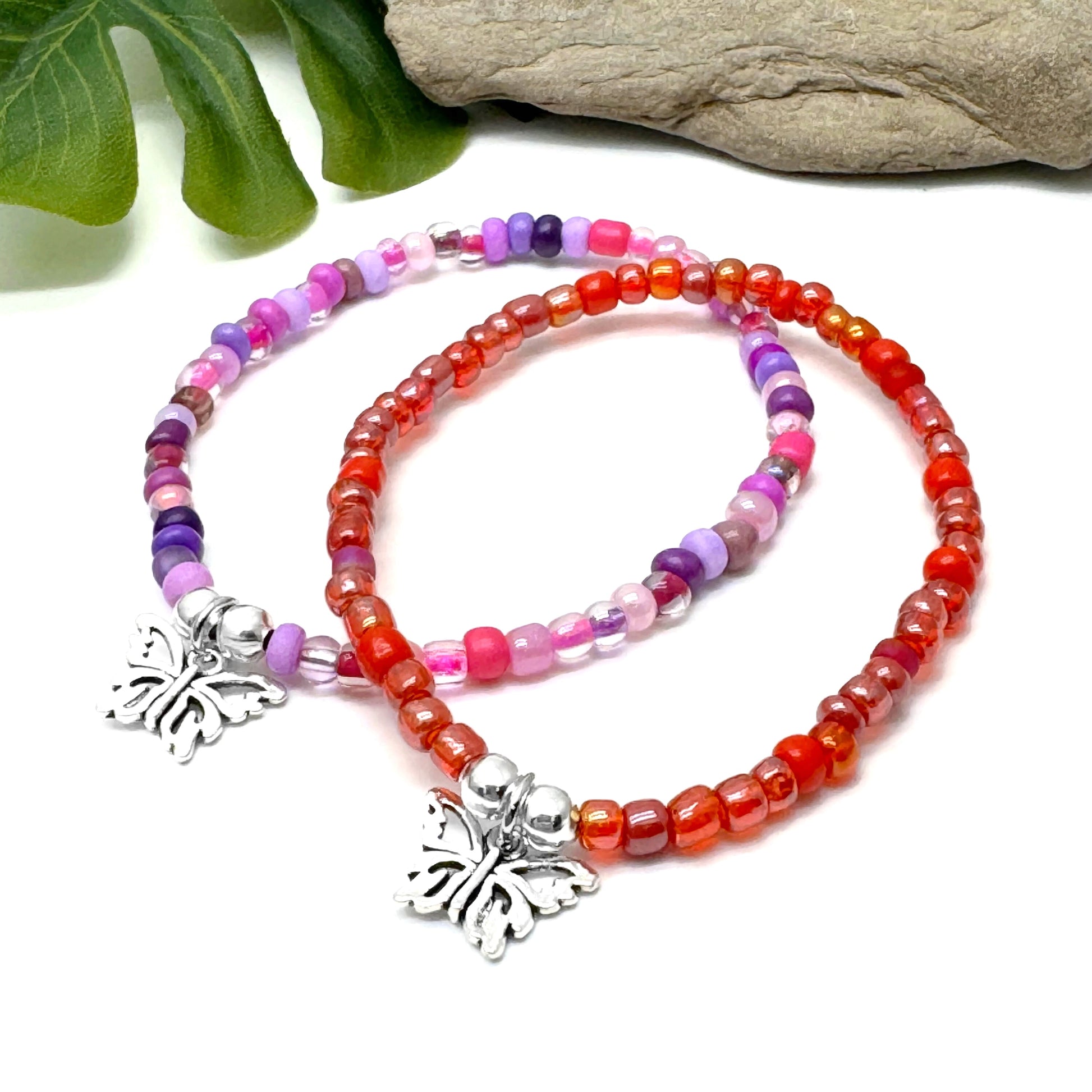 two colorful beaded bracelets with butterfly charms, placed on a white surface next to a rock.
