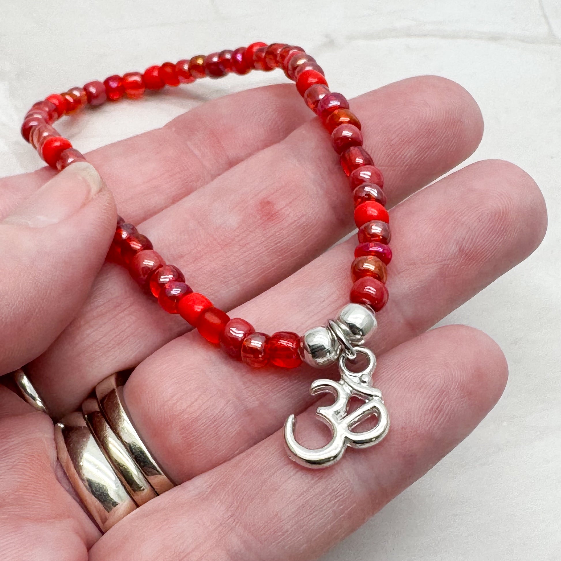 A hand holding a bracelet made of red beads with a silver charm featuring the Om symbol.