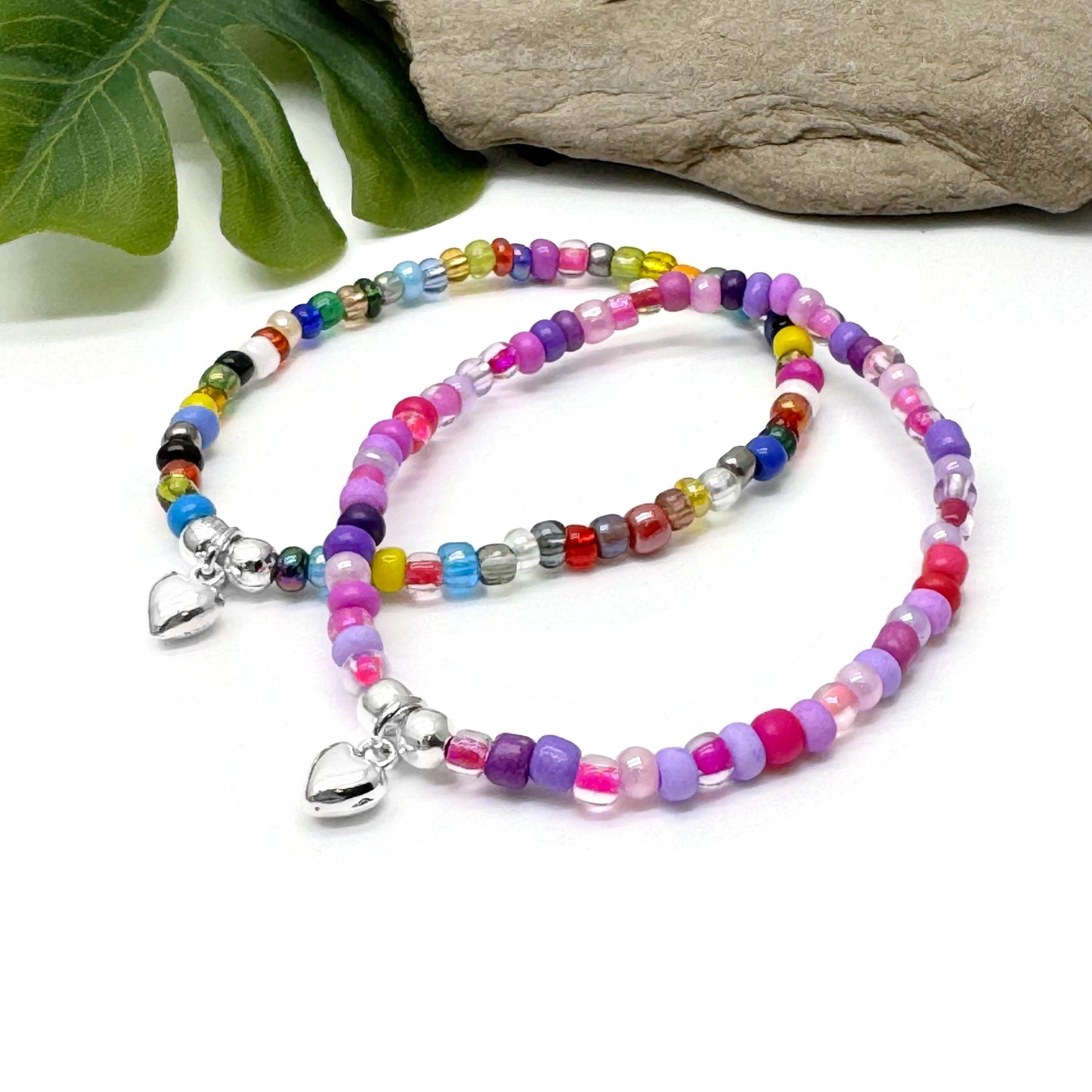 Two colorful beaded bracelets with heart-shaped charms are displayed on a white surface, accompanied by a green leaf and a rock.