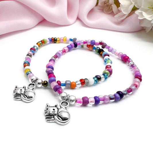 A pair of colorful beaded bracelets with cat charms, placed on a pink fabric background.