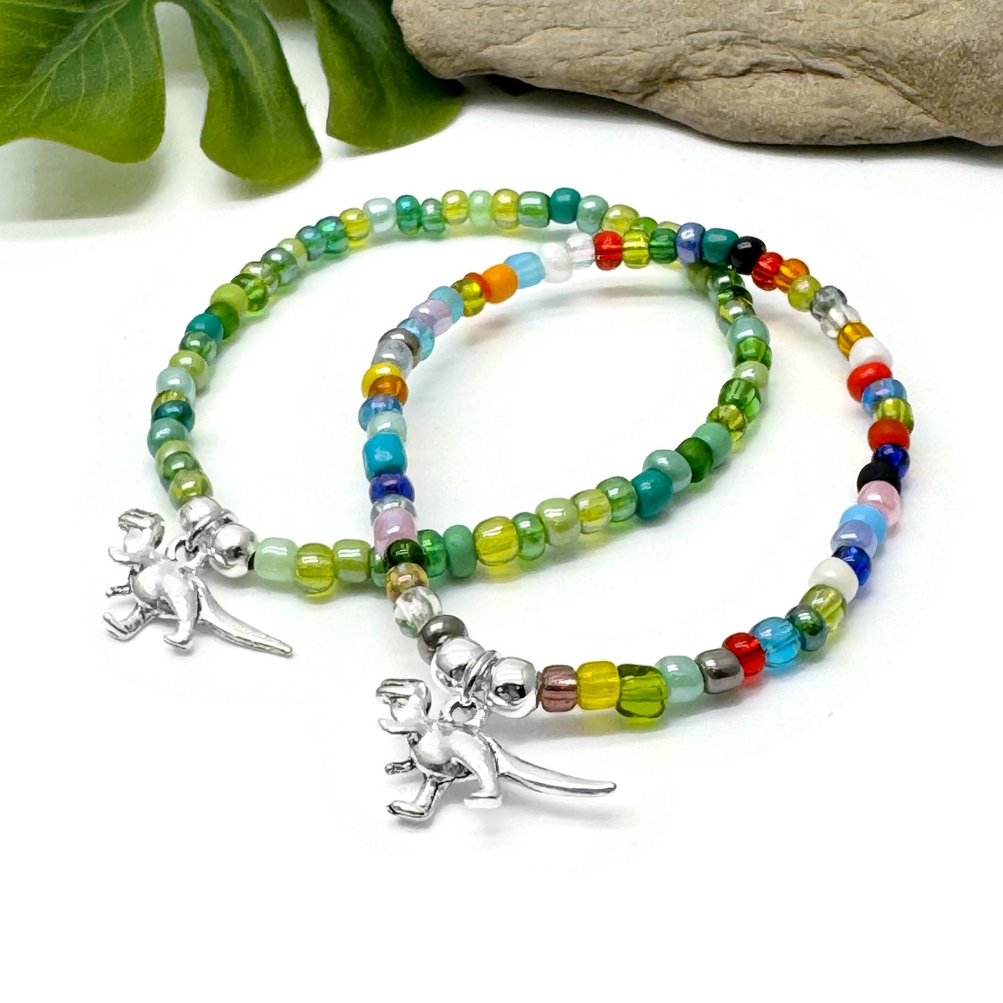 Two colorful beaded bracelets with metal charms, one shaped like a dinosaur, are displayed on a white surface.