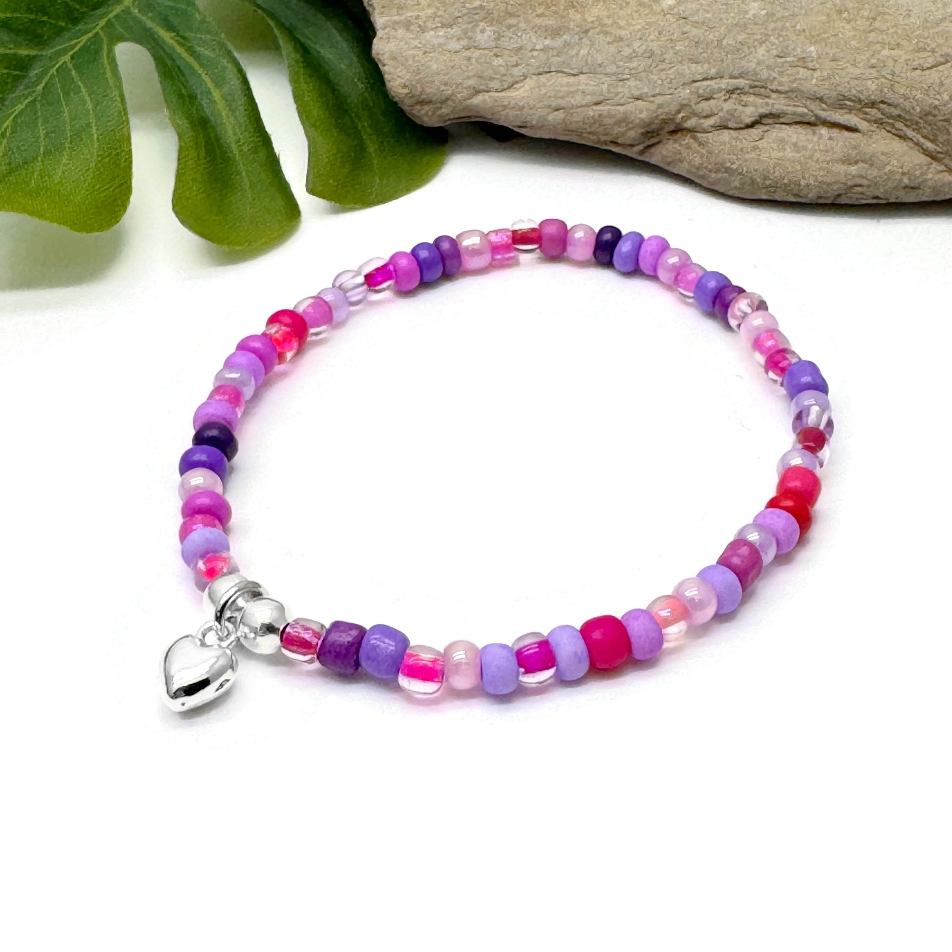 A colorful beaded bracelet with a silver charm is displayed on a white surface, accompanied by a green leaf and a rock.