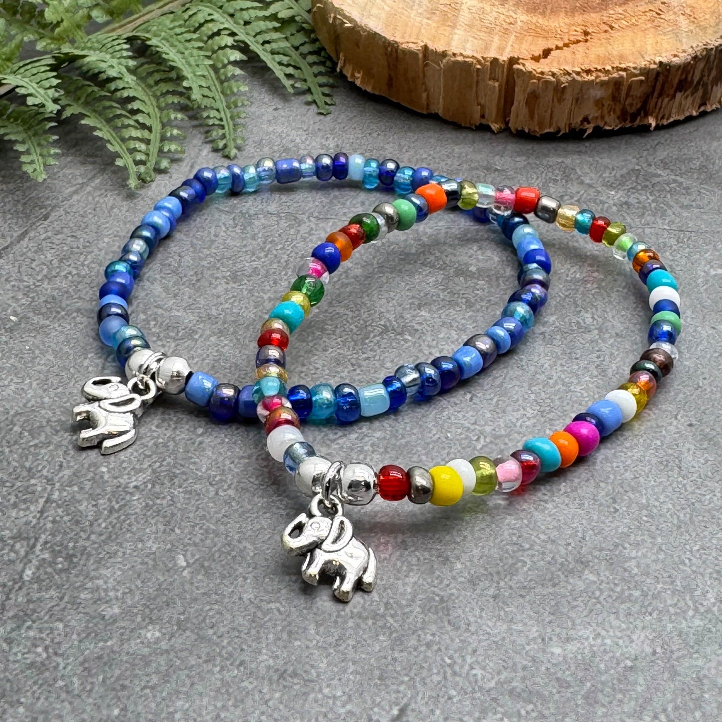two colorful beaded bracelets with elephant charms, placed on a gray surface next to a wooden log.