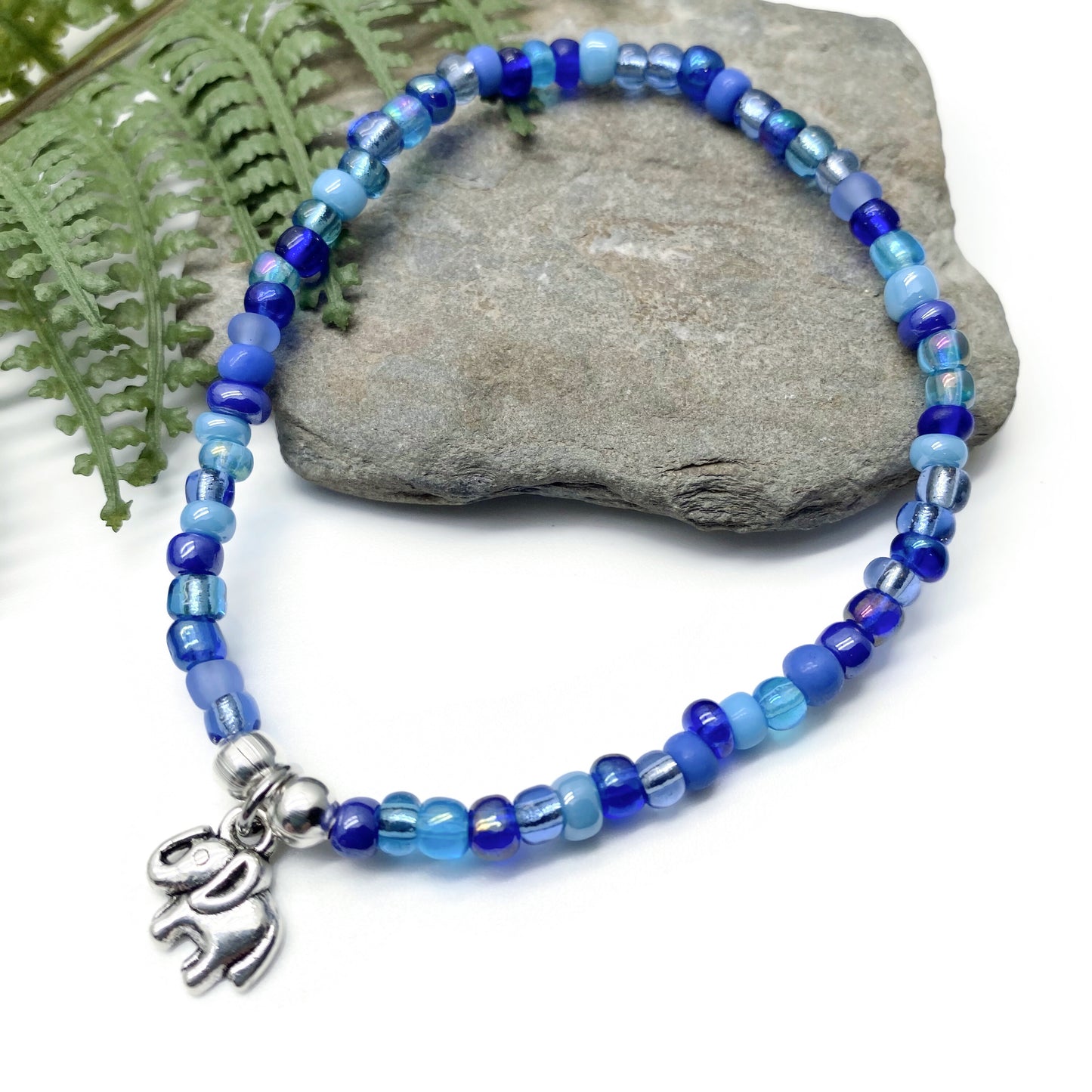 A beaded bracelet with blue and silver beads, featuring a charm in the shape of an elephant, is displayed on a rock.