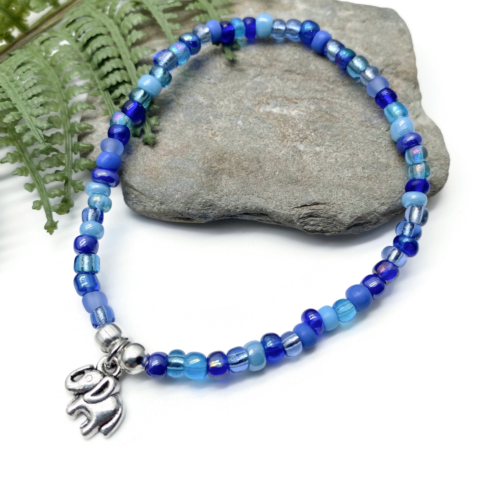 A beaded bracelet with blue and silver beads, featuring a charm in the shape of an elephant, is displayed on a rock.