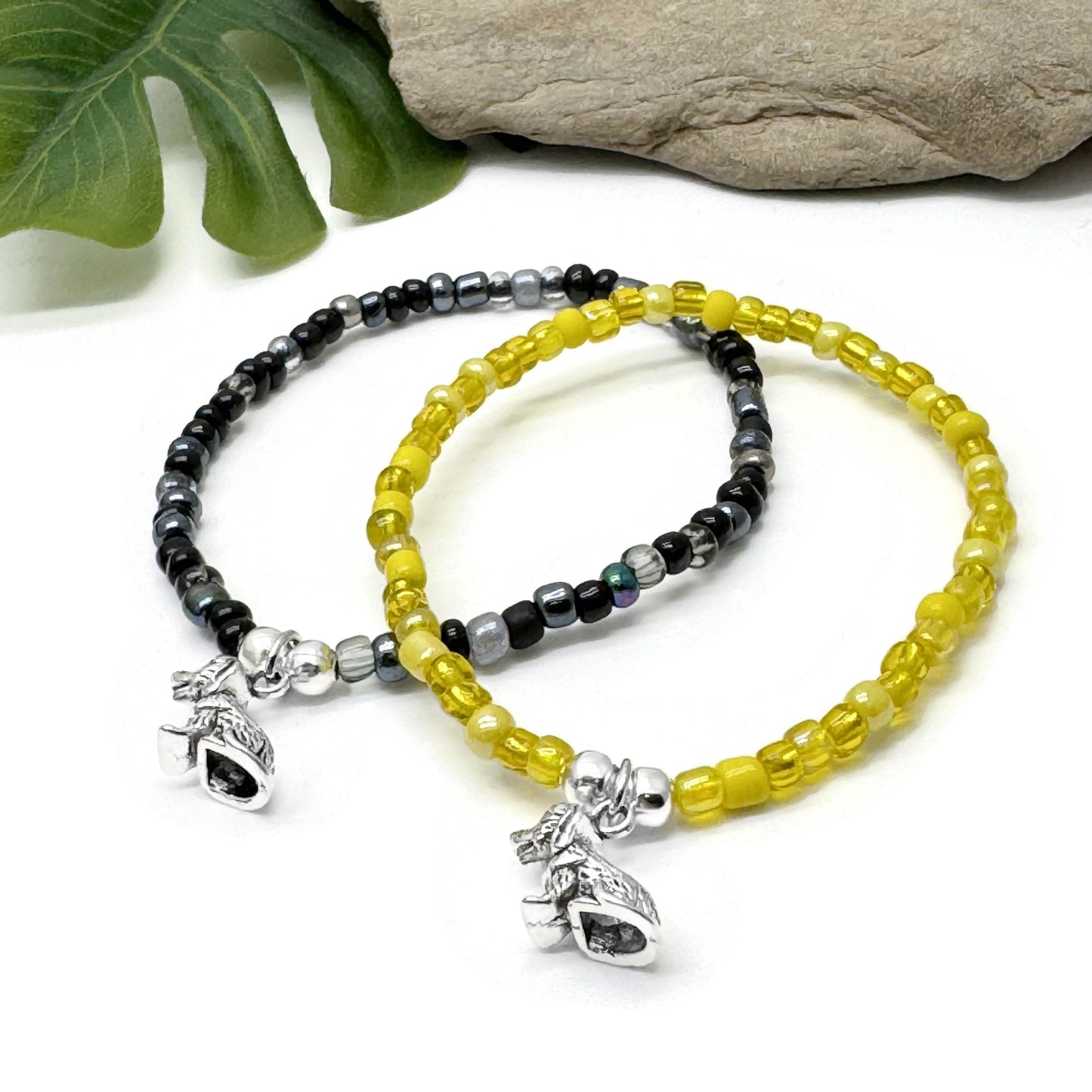 Two beaded bracelets with charms, one in black and one in yellow, are placed on a white surface with a rock and green leaves in the background.