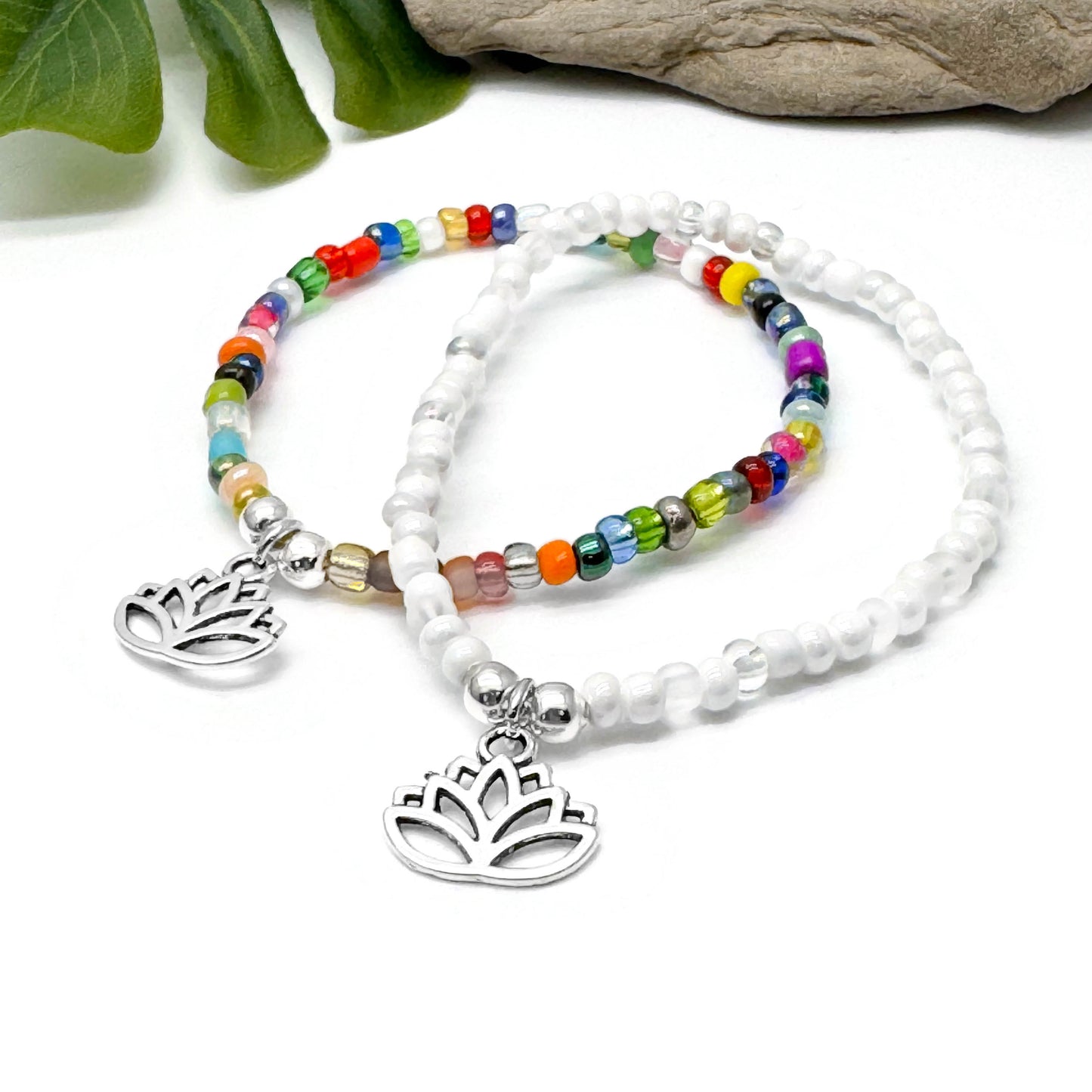 two beaded bracelets with metal charms, one with a lotus flower design, placed on a white surface with green leaves in the background.
