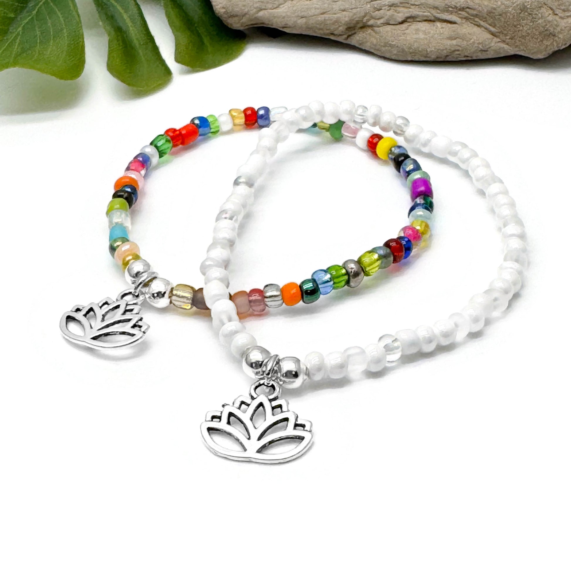 two beaded bracelets with metal charms, one with a lotus flower design, placed on a white surface with green leaves in the background.
