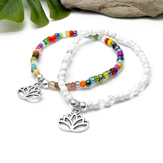 two beaded bracelets with metal charms, one with a lotus flower design, placed on a white surface with green leaves in the background.