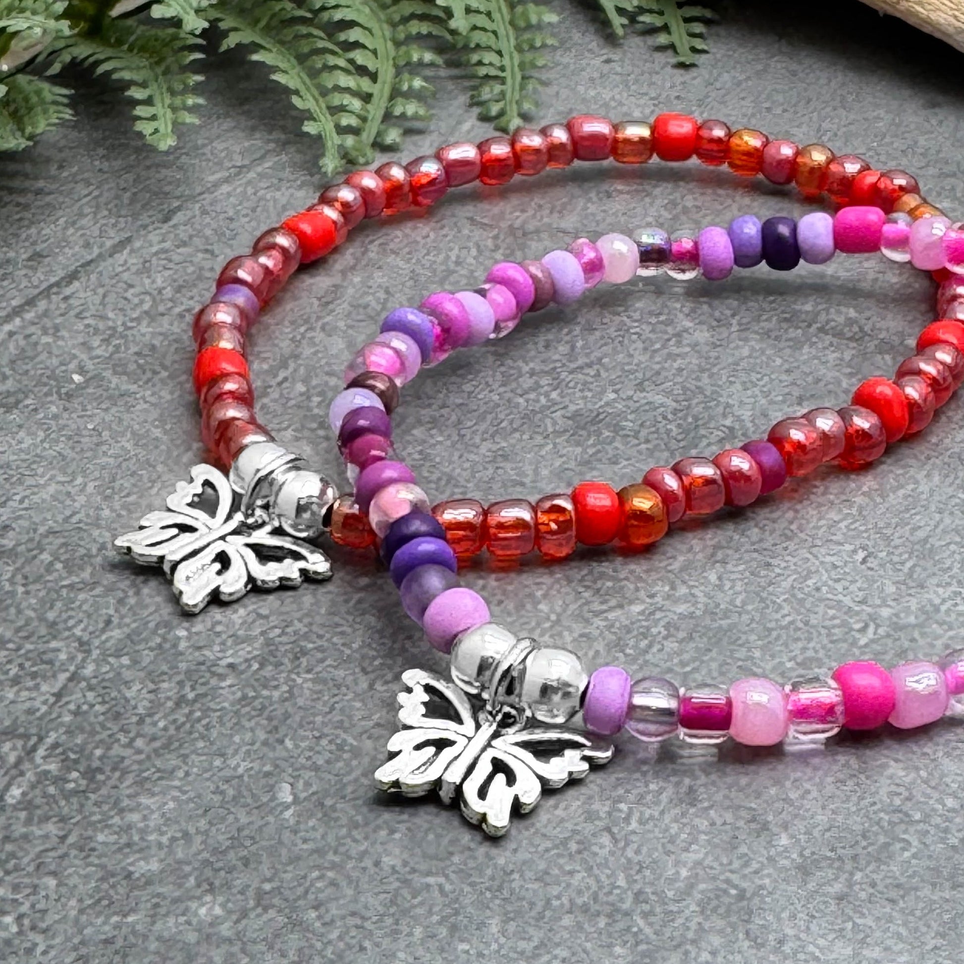 two beaded bracelets with butterfly charms, resting on a gray surface. The bracelets are adorned with a variety of colorful beads, including red, pink, purple, and clear beads, creating a vibrant and eye-catching display.