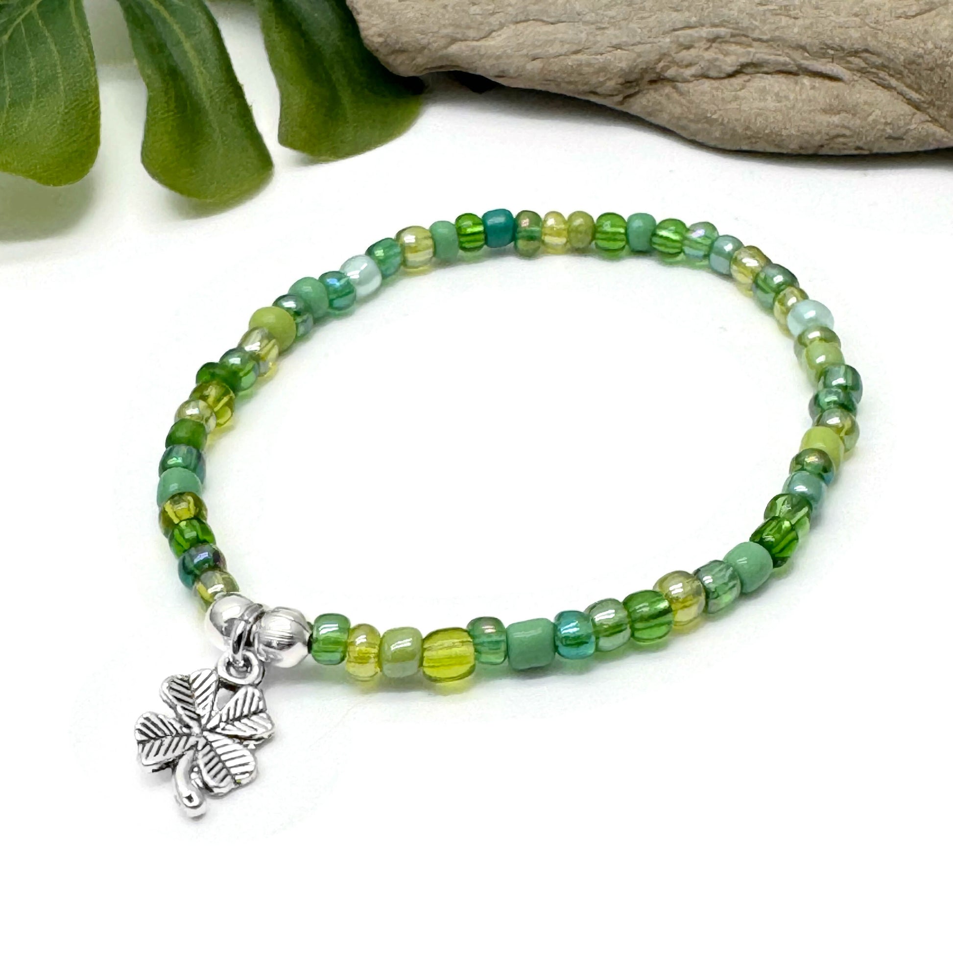 A bracelet made of green beads and a silver clover charm is displayed on a white surface.