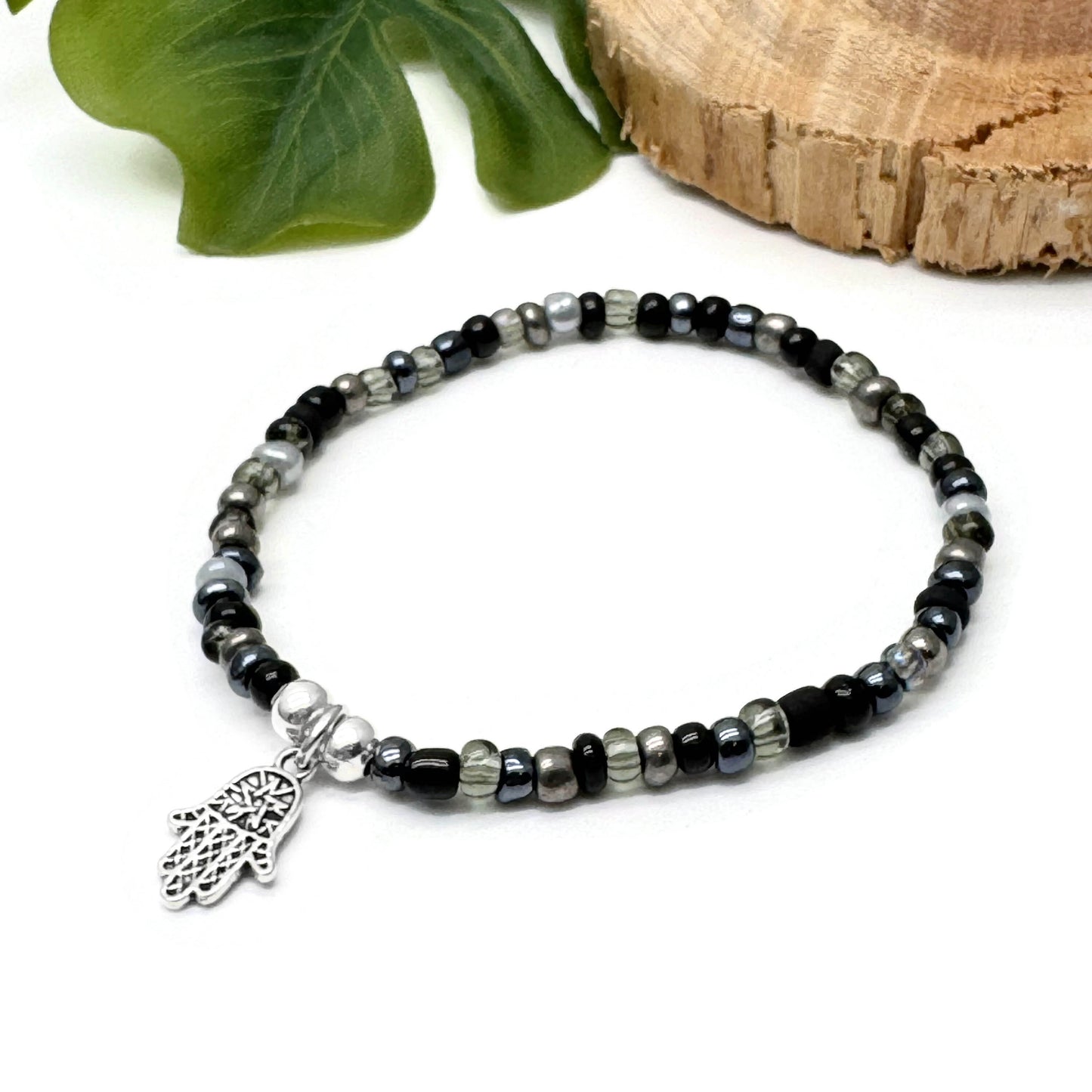 A beaded bracelet with a metal charm featuring a hand design, placed on a white surface next to a green leaf and a wooden log.