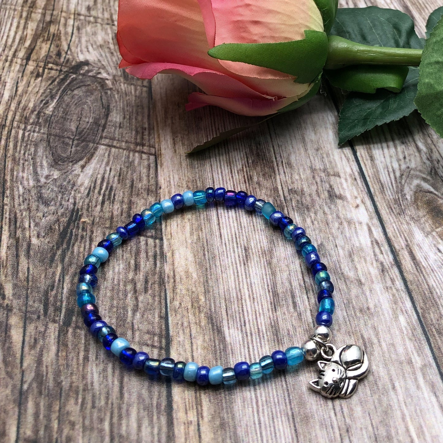 A beaded bracelet with blue and blue-green beads rests on a wooden surface, accompanied by a pink rose and a silver charm.