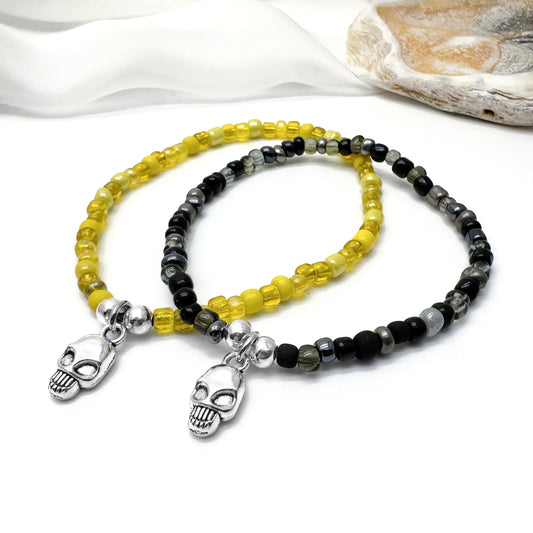two beaded bracelets with skull charms, one with yellow beads and the other with black and gray beads, placed on a white surface.