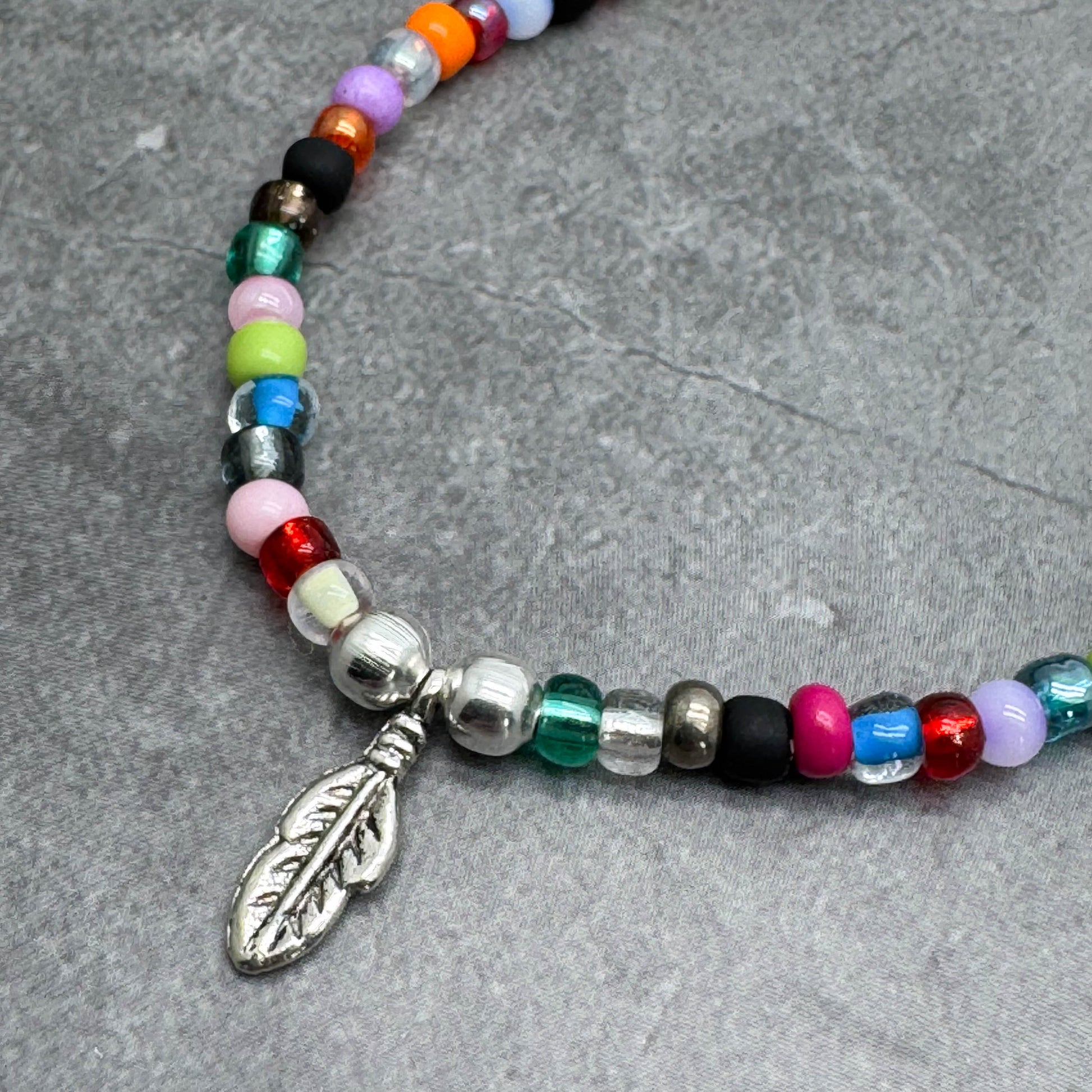 A colorful beaded necklace with a silver feather pendant is displayed on a gray surface.