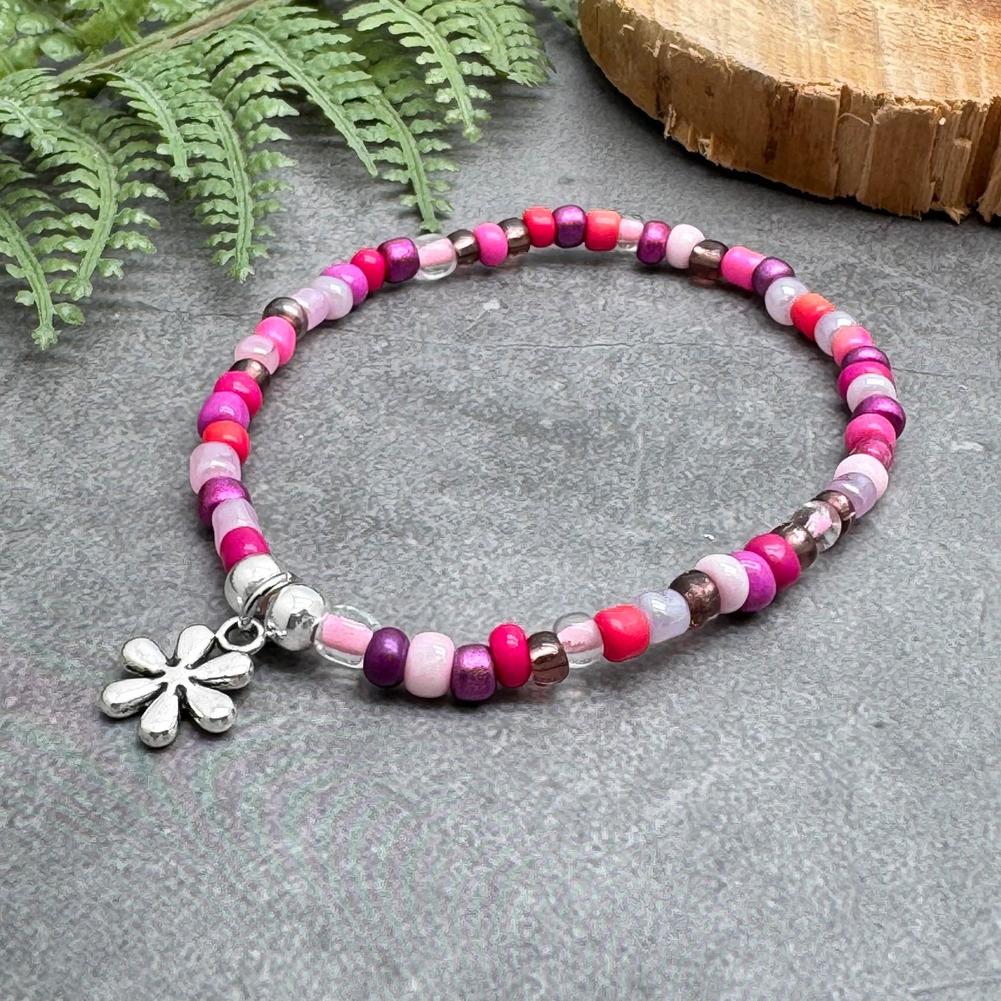 A colorful beaded bracelet with a flower charm is displayed on a gray surface, accompanied by a fern leaf and a wooden object.