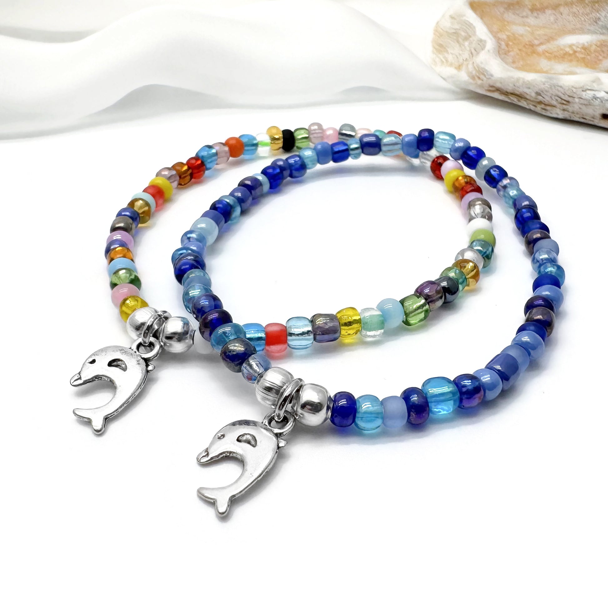 two colorful beaded bracelets with metal charms, one silver and one silver with a dolphin charm, placed on a white surface.