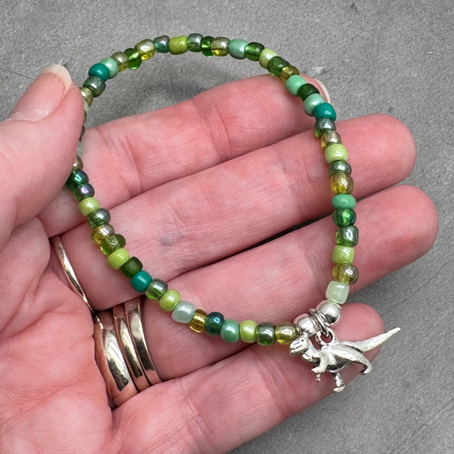 A hand holding a bracelet made of green beads and a small silver dinosaur charm.