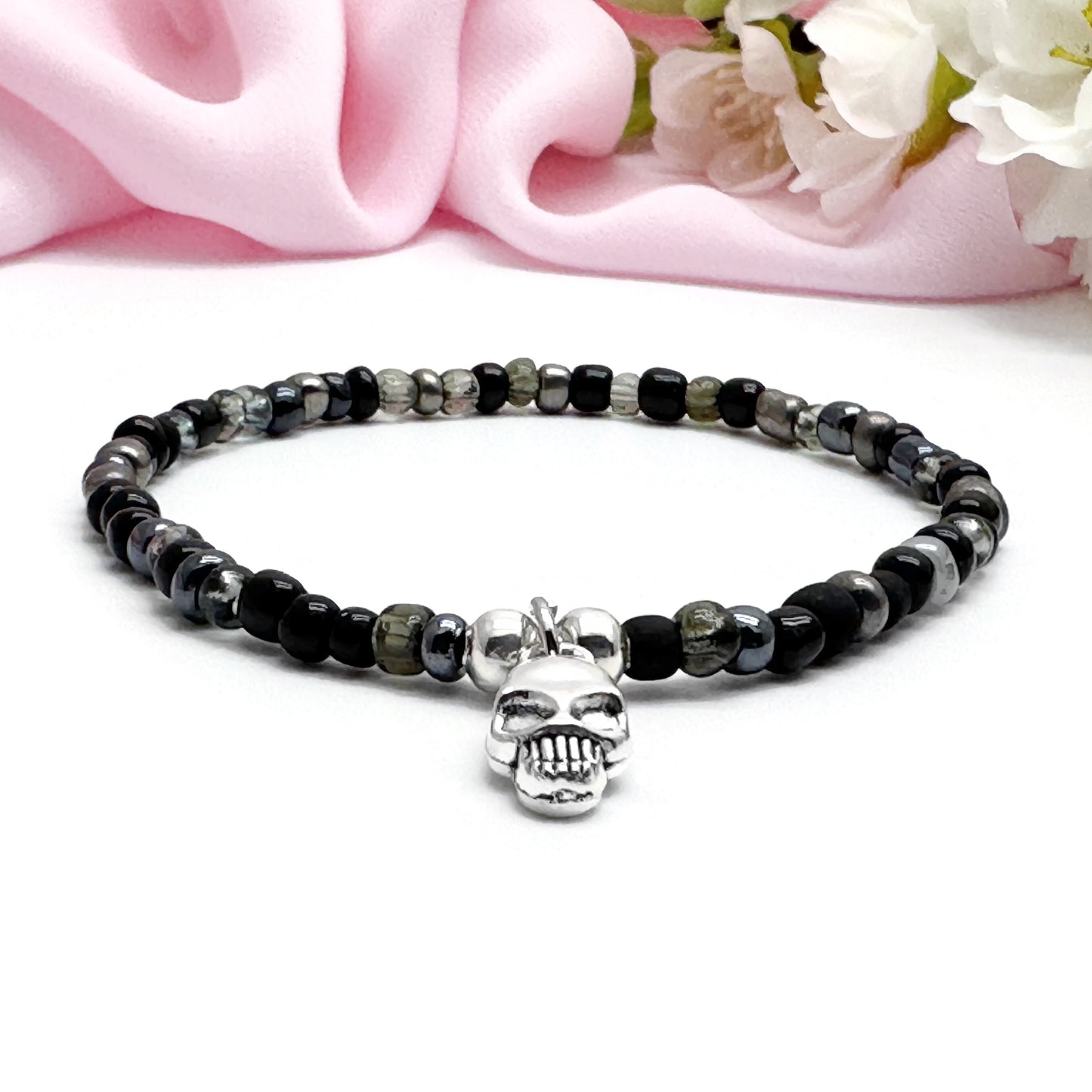 A black and silver beaded bracelet with a skull charm is displayed on a white surface, with a pink fabric in the background.