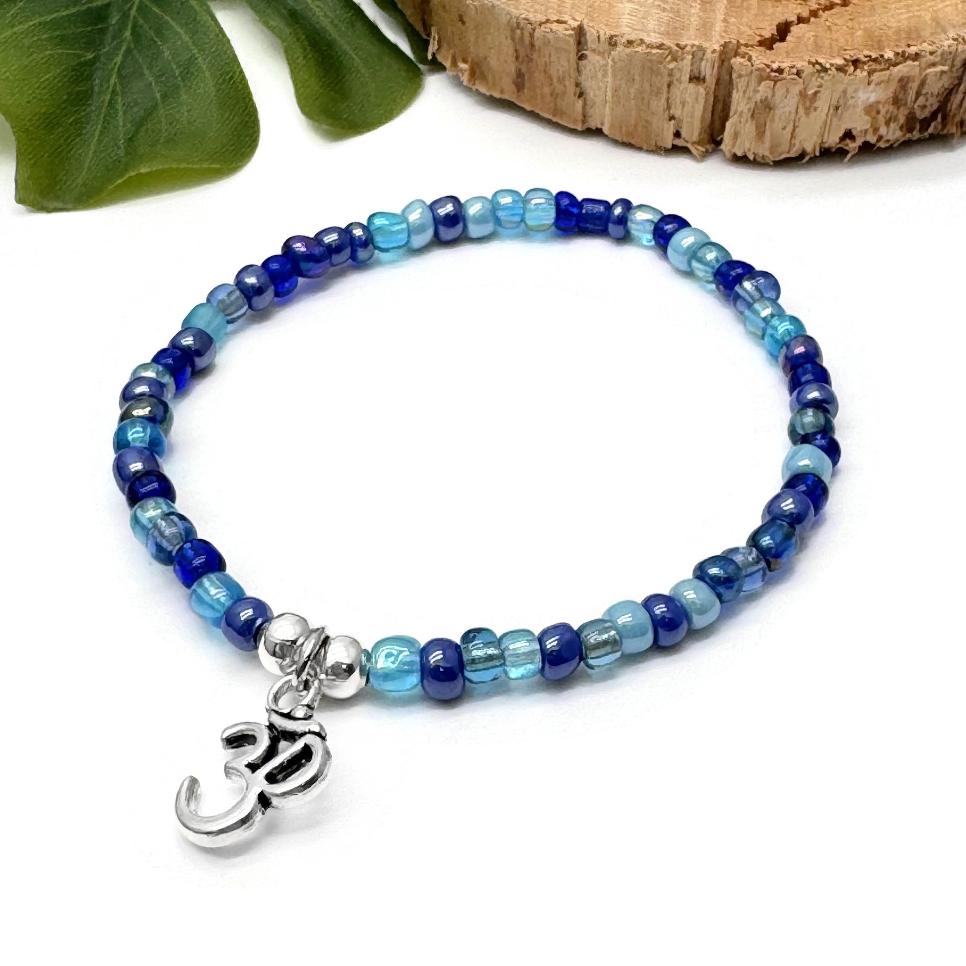 A beaded bracelet with blue and silver beads, featuring a charm with the Om symbol, placed on a white surface with green leaves in the background.
