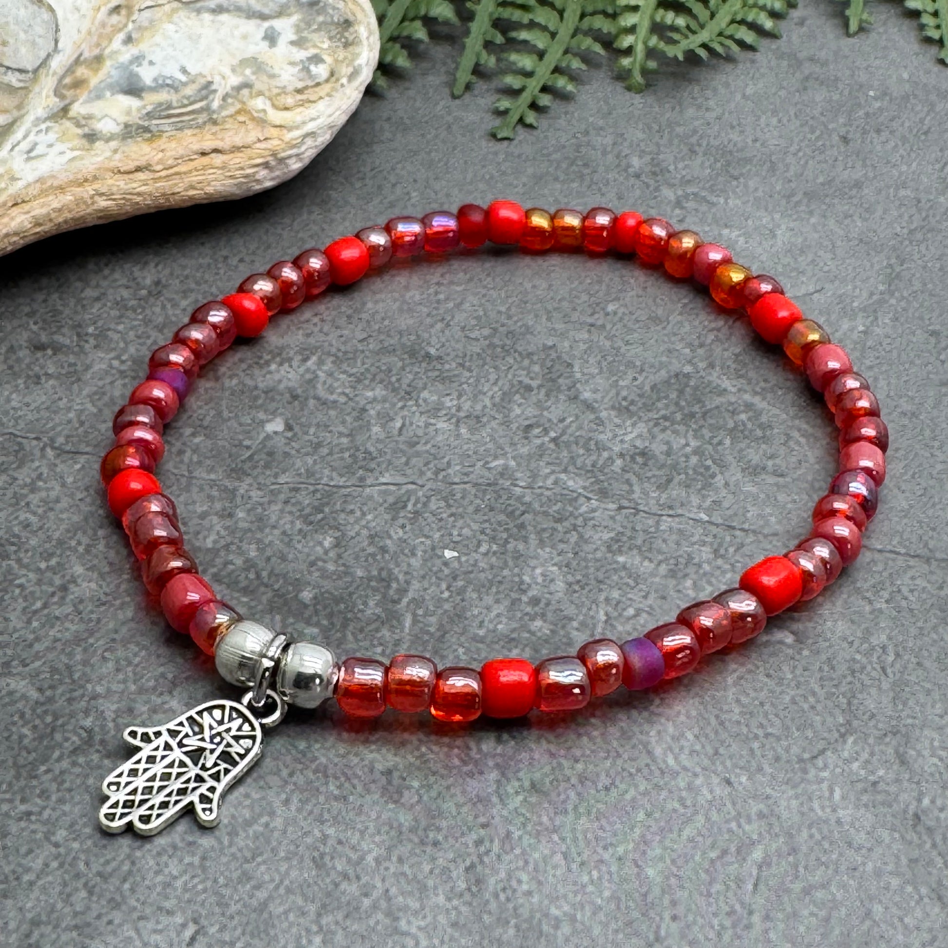 A red beaded bracelet with a silver charm is displayed on a gray surface, accompanied by a small rock and some greenery.