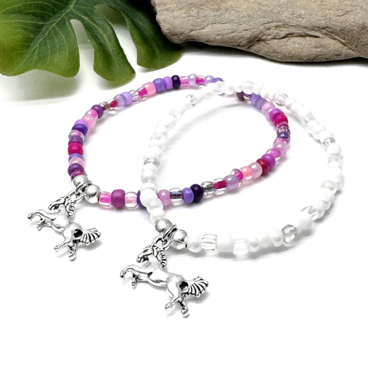 Two beaded bracelets with charms, one silver and one white, are placed on a white surface next to a green leaf and a rock.