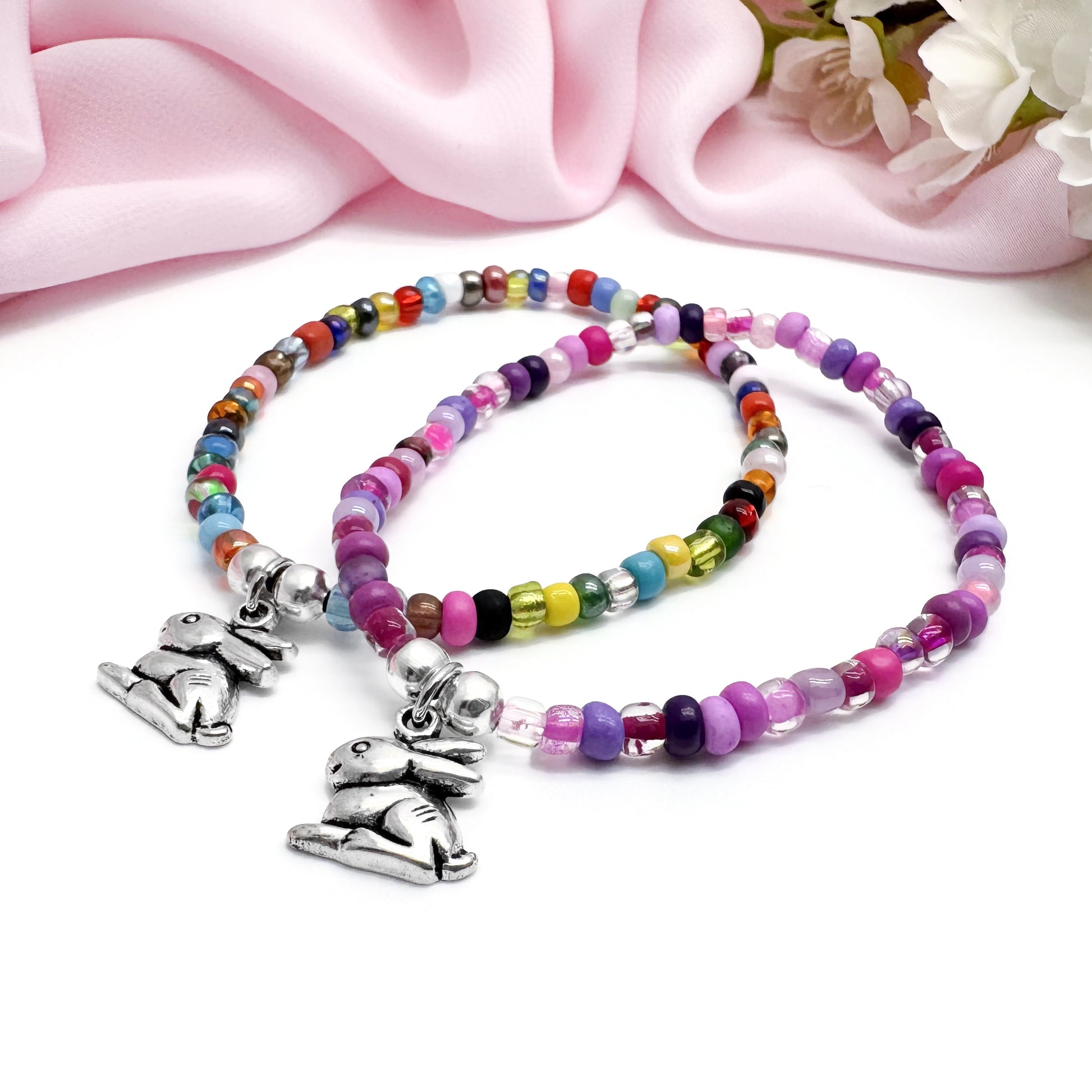 two beaded bracelets with charms, placed on a pink fabric background.